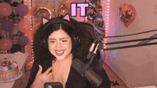 a woman wearing headphones and a cat ear headband smiles in front of a microphone with the word it above her head
