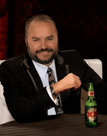 a man in a suit sits at a table with a bottle of dos equis