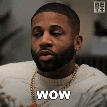 a man with a beard is wearing a white sweater and a gold chain around his neck and says wow