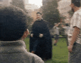 a man in a black cape is standing in a park talking to a group of people .
