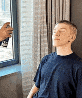 a young man is sitting in front of a window with his eyes closed while a person sprays a spray on his face .