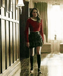a woman is walking down a hallway wearing a red sweater and a plaid skirt