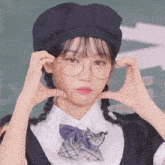 a girl wearing glasses and a hat makes a heart with her hands