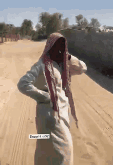 a man with a scarf around his head is standing on a dirt road with snaph ad2 on the bottom right