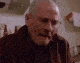 a bald man wearing glasses and a brown jacket is sitting at a table in a kitchen .
