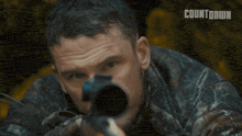 a man in a camo jacket is aiming a sniper rifle with a countdown logo in the background