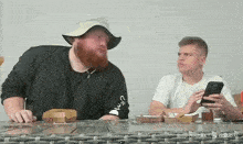 a man with a beard is sitting at a table while another man looks at his phone .