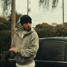 a man wearing a beanie and a hoodie stands in front of a car