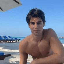 a shirtless man sits on a beach with a blue chair in the background