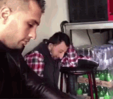two men are sitting on stools in a room with bottles of soda on the shelf .