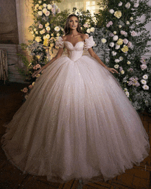 a woman in a white ball gown stands in front of a wall of flowers