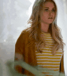 a woman in a yellow and white striped shirt and a brown cardigan is standing in front of a window .
