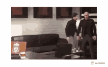two men are standing in a living room next to a couch and a table .