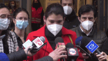 a woman wearing a red jacket is surrounded by microphones including one that says ' agencia uno '