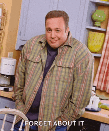 a man in a plaid shirt is standing in a kitchen with the words " forget about it " on the bottom