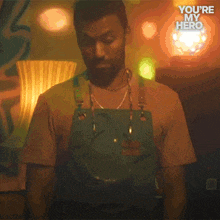 a man wearing an apron is standing in a dark room with the words " you 're my hero " written above him