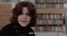 a woman is sitting in a library with a bookshelf in the background and says `` eat shit '' .