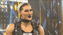 a female wrestler wearing a black vest with spikes and a choker looks at the camera