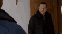 a man in a black jacket stands in front of a door with uc on the bottom right