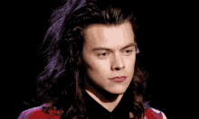 harry styles is wearing a red jacket and a black shirt with long hair and pink lipstick .