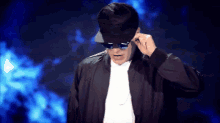 a man wearing a hat and sunglasses adjusts his sunglasses against a blue background