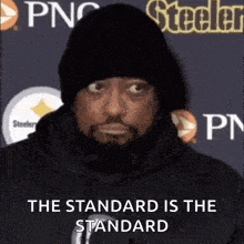 a man with a beard wearing a beanie and a hoodie is standing in front of a steelers logo .