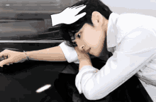 a man in a white shirt is leaning on a black car with a hand on his forehead