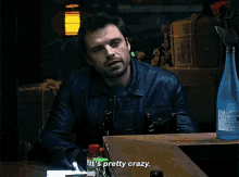 a man in a leather jacket is sitting at a table with a bottle of water and says it 's pretty crazy .