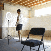 a man in a white shirt stands in front of a ring light and a folding chair