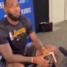 lebron james is sitting in a chair holding a book while wearing a lakers shirt .