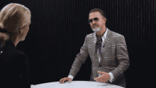 a man wearing sunglasses and a plaid jacket talks to a woman
