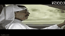 a man in a white robe is driving a car with the word kooo3 on the bottom right