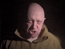 a bald man wearing a brown jacket looks at the camera