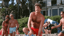 a shirtless man in red shorts with a lifeguard logo on his shorts sits on the beach