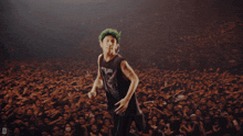 a man with green hair is singing into a microphone in front of a crowd with a staff shirt in the background