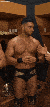 a shirtless wrestler in a locker room talking to another wrestler with a crown on his knee pads