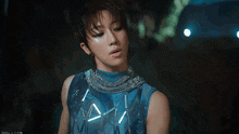 a man wearing a blue sleeveless top with a necklace around his neck is standing in a dark room
