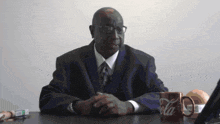 a man in a suit sits at a desk with a coca cola mug on the table