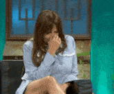 a woman in a blue shirt is sitting on a couch covering her face with her hand .
