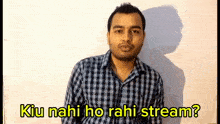 a man in a plaid shirt is standing in front of a white wall and says " kiu nahi ho rahi stream ? "