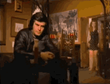 a man in a leather jacket sits in a chair in a room with a woman standing in the background