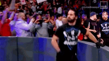 a man in a shield shirt is walking through a wrestling ring while a crowd watches .