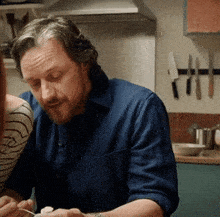 a man in a blue shirt sits in a kitchen with knives on the wall behind him