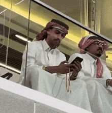 two men are sitting on a balcony and one is looking at his phone