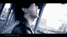 a man in a black jacket is singing into a microphone while looking out a window