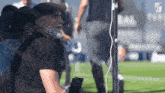 a man with a beard wearing a black shirt and a hat stands on a soccer field
