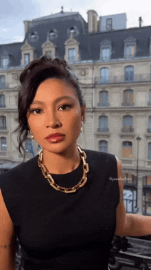 a woman wearing a black dress and a gold chain necklace is standing in front of a building .