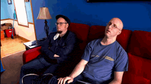 two men sitting on a red couch with one wearing a blue shirt that says ' columbia ' on it