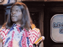 a man with long hair is holding a microphone in front of a sign that says uh-e