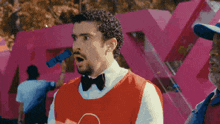 a man in a red vest and bow tie is standing in front of a large pink x